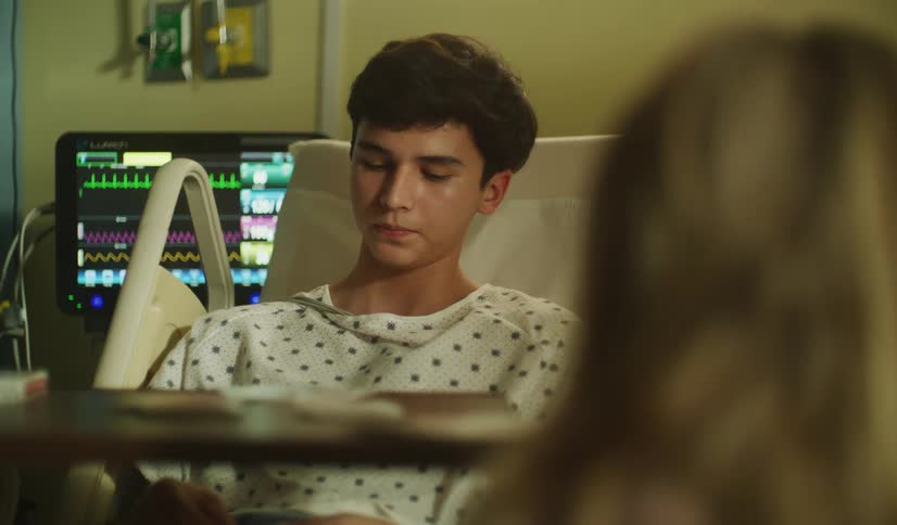A teenage boy in a hospital gown smiles and then grows serious while talking with a visitor in his hospital room