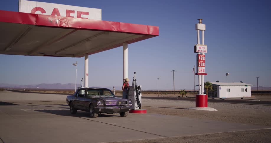 A woman in a cowboy hat stops her classic muscle car to refuel at a retro roadside gas station and cafe in the sunny desert