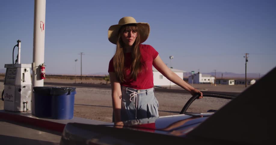 A young woman wearing a sun hat patiently waits while refueling her vintage car at a secluded desert gas station