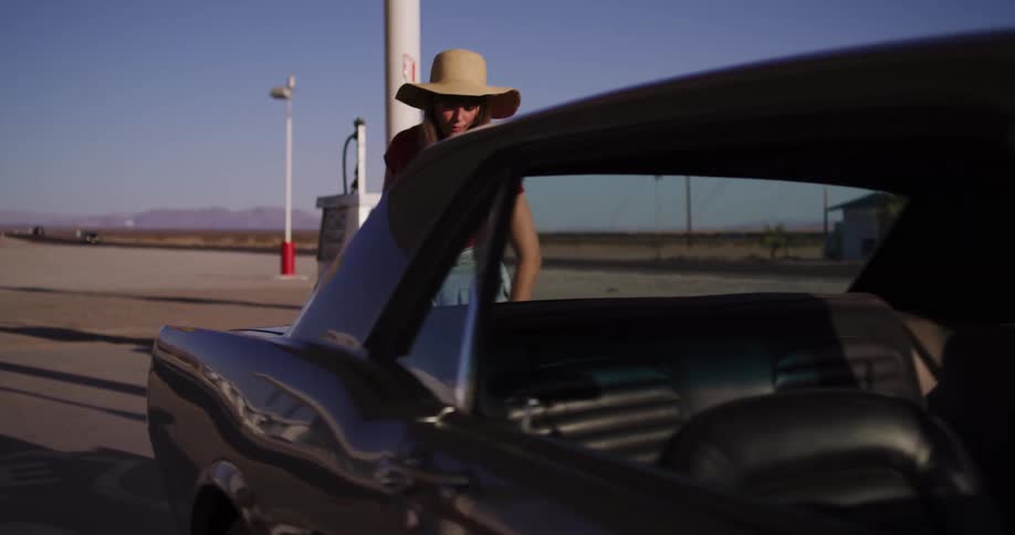 A stylish woman in a hat refuels her classic car at a remote desert gas station on a sunny day