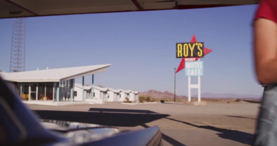 A woman finishes fueling her car at a desolate desert gas station with a classic Roy's Motel Cafe sign in the background
