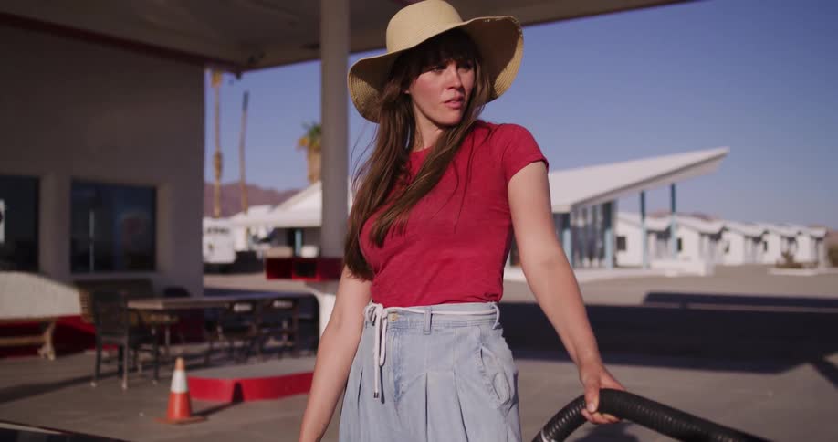 A woman in a red shirt and straw hat refuels her car in slow motion at a vintage desert gas station