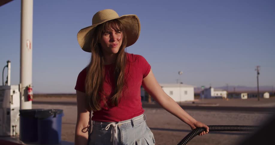 A woman in a straw hat stands at a gas pump before the camera pulls back to reveal the iconic Roy's Motel Cafe sign in the California desert