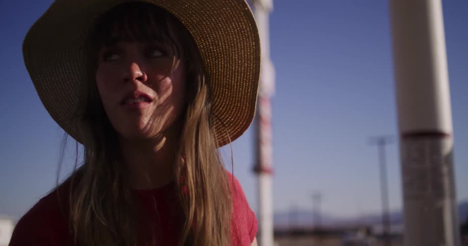 A cinematic slow-motion close-up of a young woman in a floppy straw hat with a pensive expression at a roadside gas station