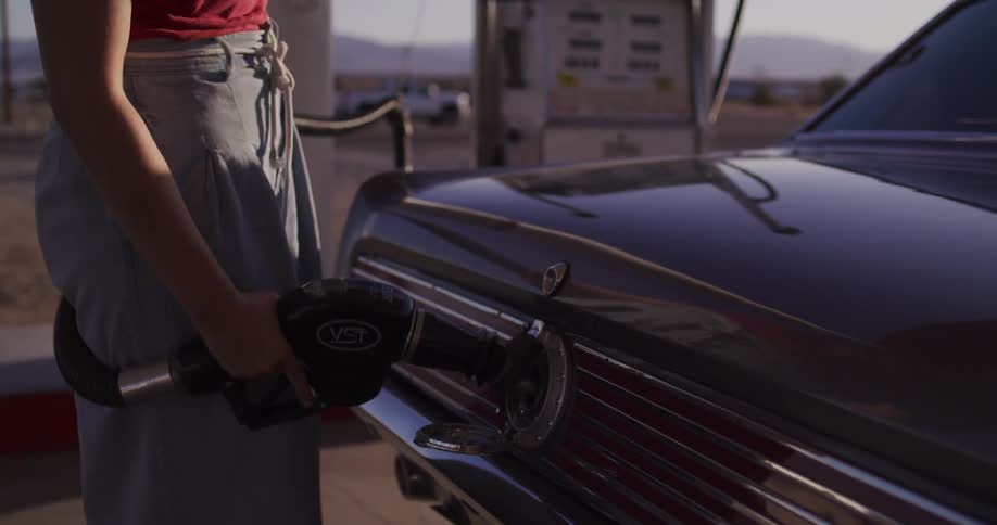 A young woman in a hat gets out of her classic car to pump gas at a deserted station in the sunny desert