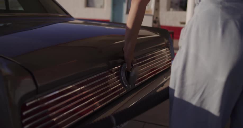 A stylish young woman refuels her vintage car at a remote roadside gas station in the desert sun