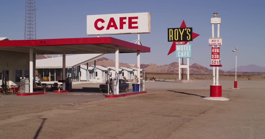 A woman driving a classic black muscle car pulls into Roy's Motel and Cafe gas station in the desert to refuel