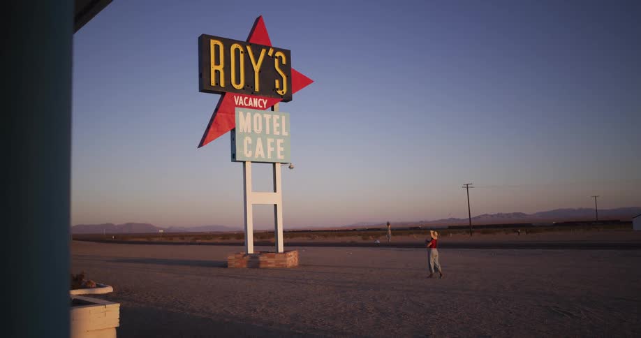 A lone woman in vintage attire stands under the iconic Roy's Motel Cafe sign on a desolate desert highway at sunset