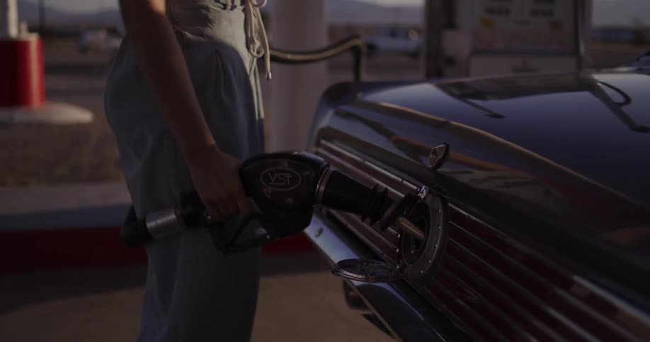 A woman in a straw hat pumps gasoline into her vintage car at a remote desert gas station during a beautiful sunset