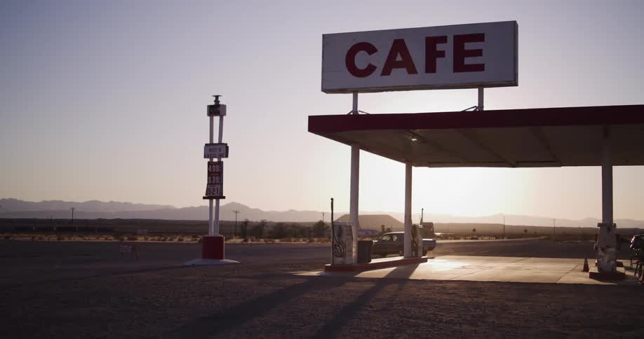 A classic car drives away from a remote gas station and cafe in the desert at sunset