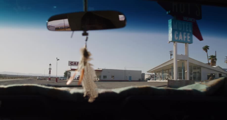A slow-motion point-of-view shot from inside a car driving towards the iconic Roy's Motel and Cafe in the desert