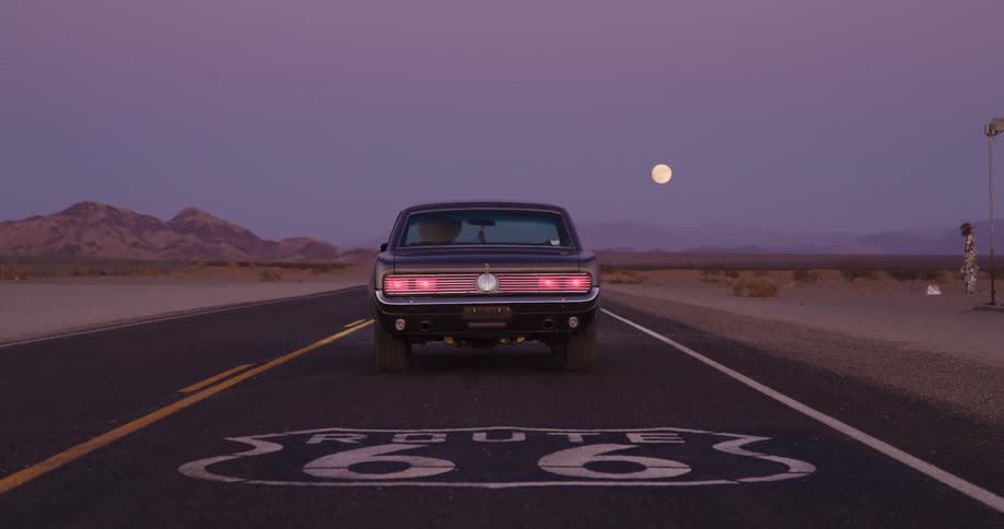 A vintage black muscle car drives away down historic Route 66 in the desert as the full moon rises at twilight