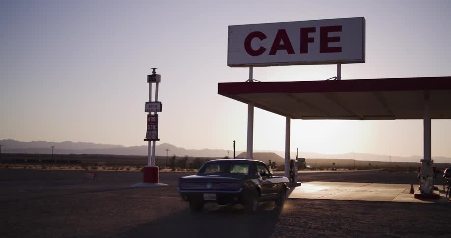A classic muscle car pulls up to the pumps at an isolated desert gas station and cafe at sunset
