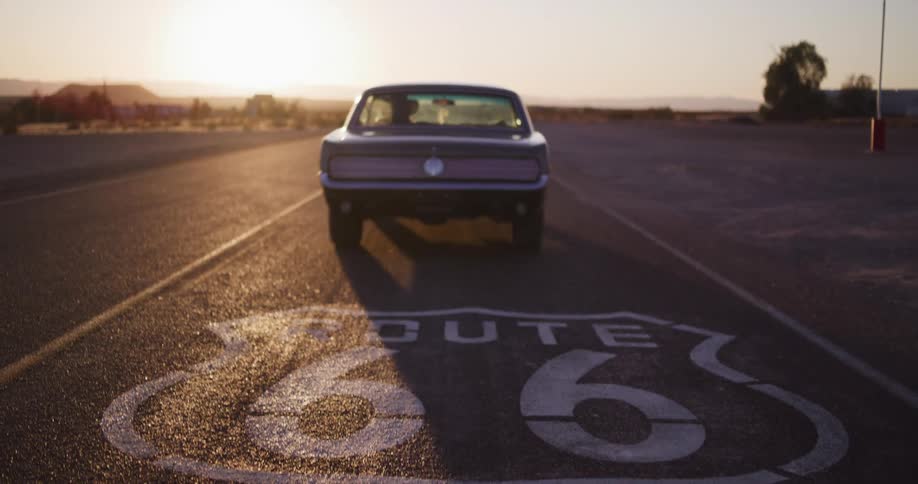 A woman in a classic car drives away from the camera on Route 66 into the desert sunset, revealing the iconic road sign
