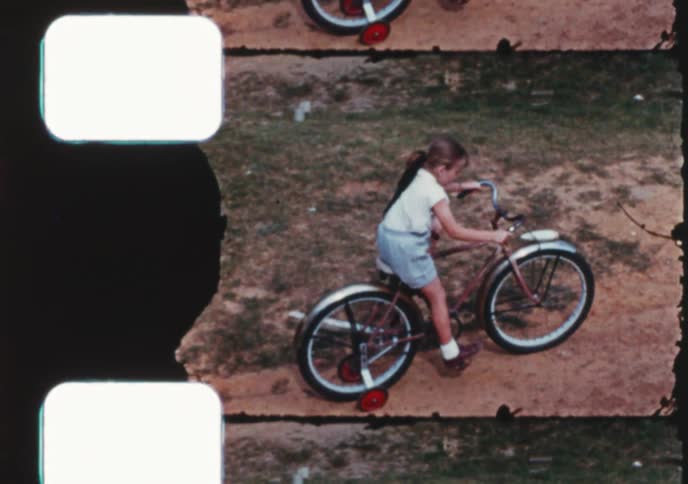 A high-angle vintage shot shows a young girl learning to ride her bicycle with training wheels in a grassy yard