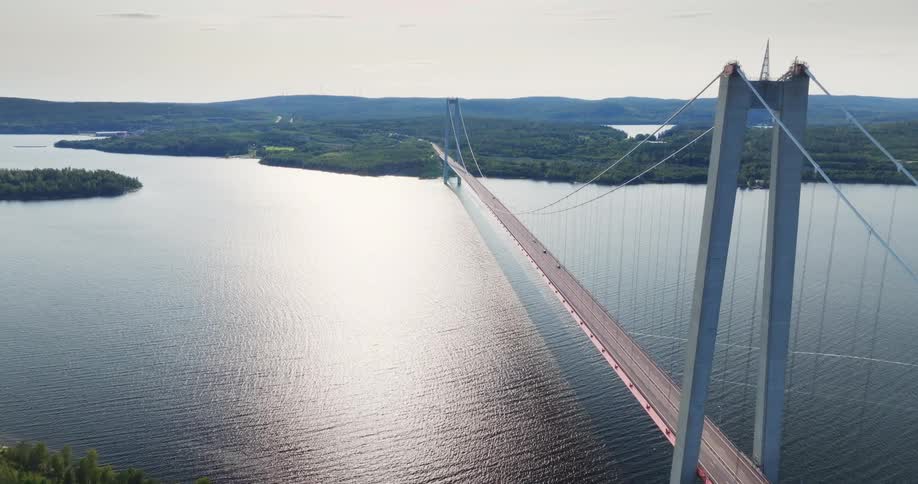 A stunning aerial view of a large suspension bridge spanning a wide river, surrounded by lush green forests under a bright sky