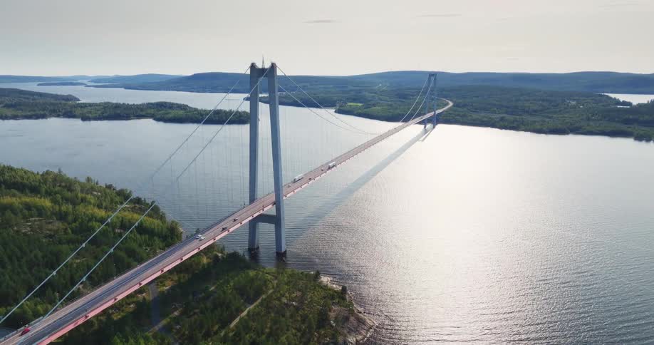 A stunning aerial shot pulls back to reveal the impressive scale of the High Coast Bridge in Sweden, spanning a vast waterway amidst a forested landscape