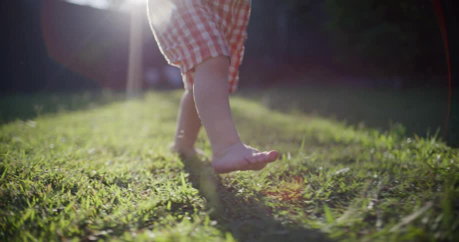 A toddler's bare feet take their first tentative steps across a sunlit green lawn in slow motion