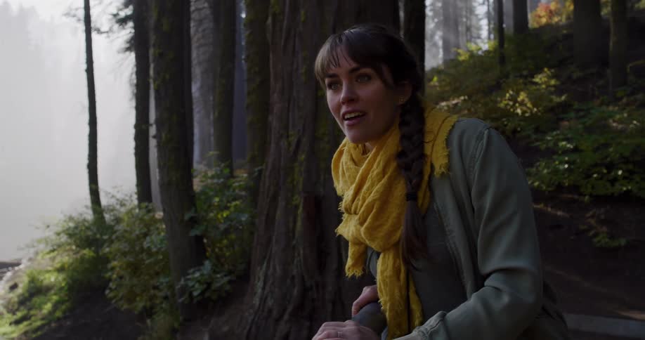 A young woman wearing a yellow scarf stops on a forest path and looks out in awe at the misty, sunlit scenery in slow motion