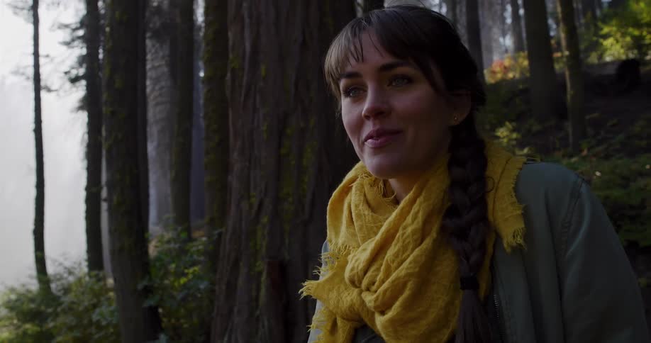A beautiful woman wearing a yellow scarf reacts with joyful wonder to an amazing sight while exploring a sunlit forest
