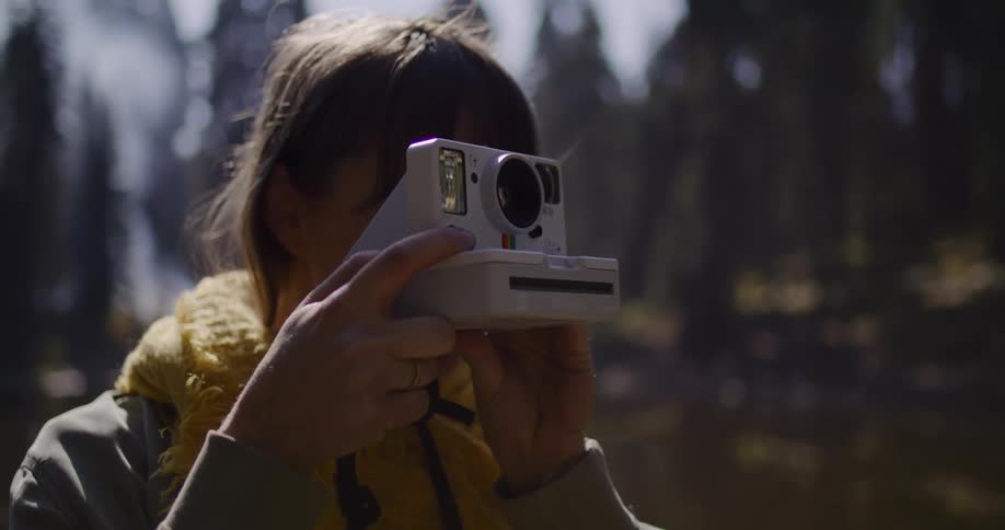 A young woman uses a vintage-style instant camera to take a photo while exploring a sunlit forest