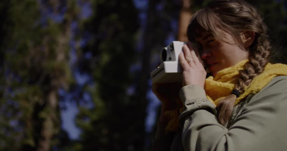 A woman touches a giant tree stump then takes a picture with her vintage instant camera in a sunny forest