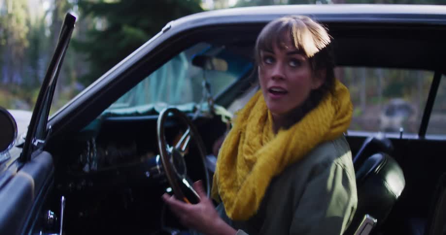 A young woman ties her hiking boots and excitedly gets into her classic car with a camera, ready for an adventure
