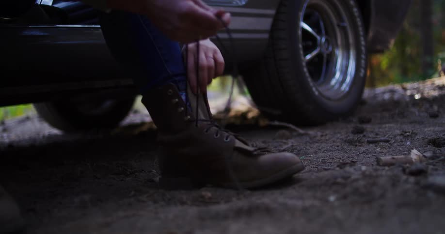 A woman sits on the side of a vintage car in the forest and laces up her leather hiking boots in slow motion