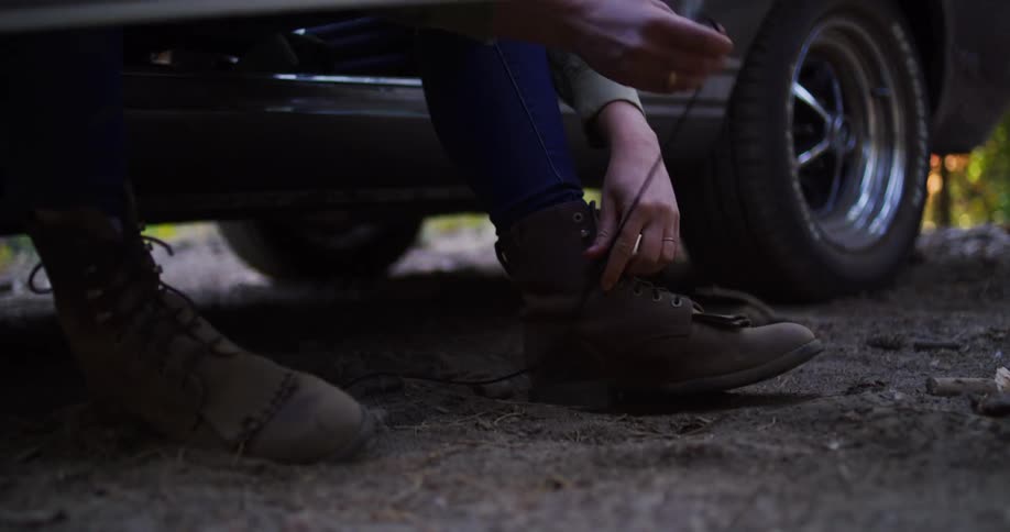 A person ties their boots while sitting in the open door of a classic car parked in a forest
