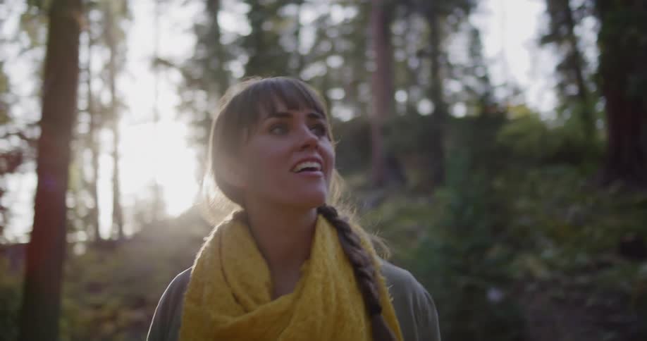 A happy young woman wearing a yellow scarf looks up in awe at the beauty of the sunlit forest around her