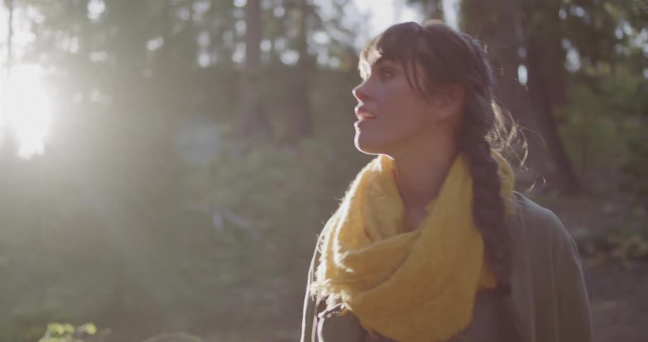 A young woman with a braided hairstyle smiles with wonder as she walks through a beautiful forest with golden sunlight streaming through the trees