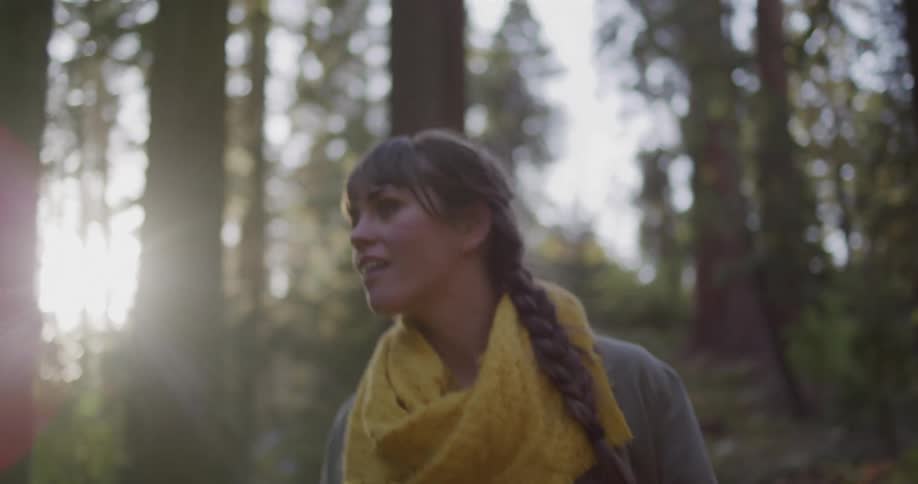 A happy young woman with a braided hairstyle and yellow scarf looks up in awe while exploring a sun-dappled redwood forest