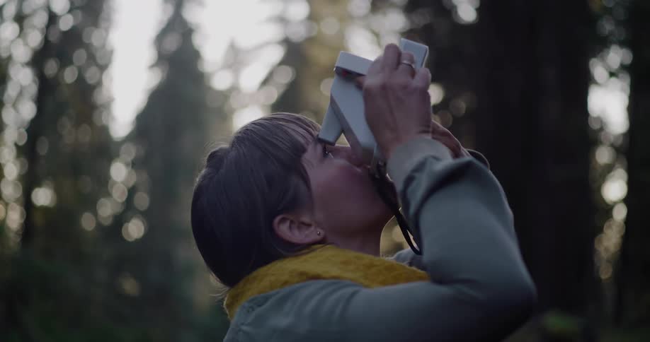 A woman with a yellow scarf gazes up in wonder at the forest canopy before taking a picture with her instant camera