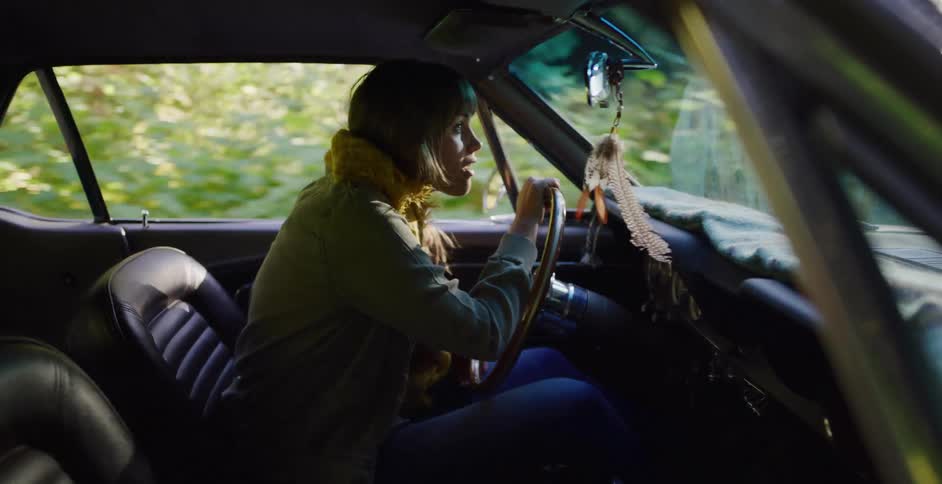 A woman in a yellow scarf drives her classic car down a road through a sun-dappled forest