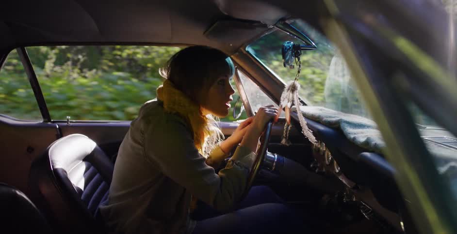 A young woman with a yellow scarf drives a classic car through a sunlit forest, her happy expression changing to one of fear as she looks back