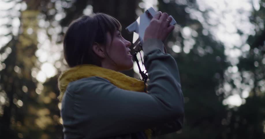 A woman gently touches the bark of a large tree in the forest and then takes a picture of the canopy with her instant camera