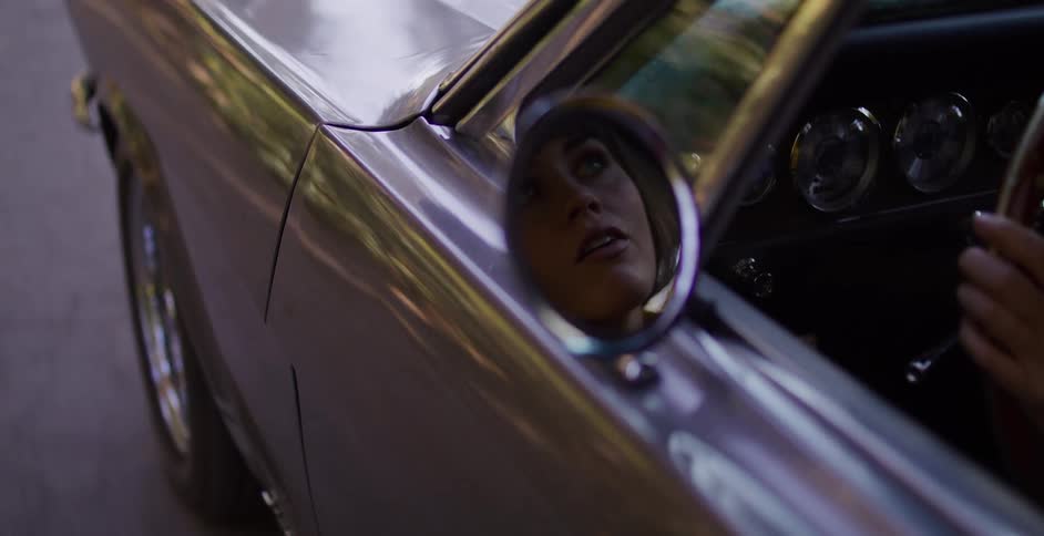 A stylish woman's face is seen in the reflection of a classic silver car's side-view mirror as she drives