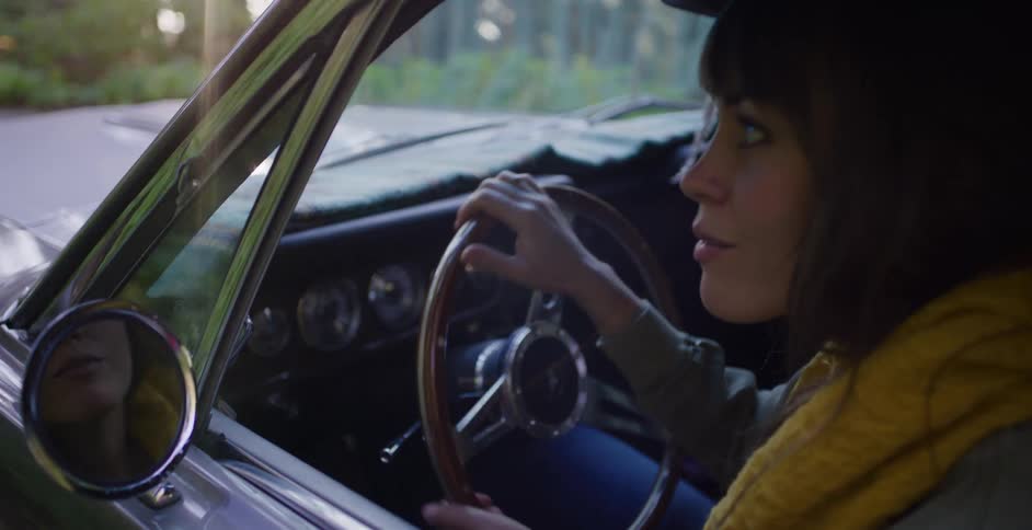 A young woman in a classic convertible car sits behind the steering wheel while parked on a country road