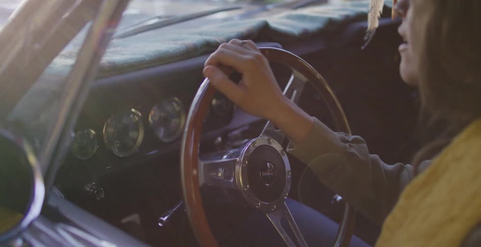 A woman's hands gently steer a classic car down a sun-dappled road during a golden hour drive