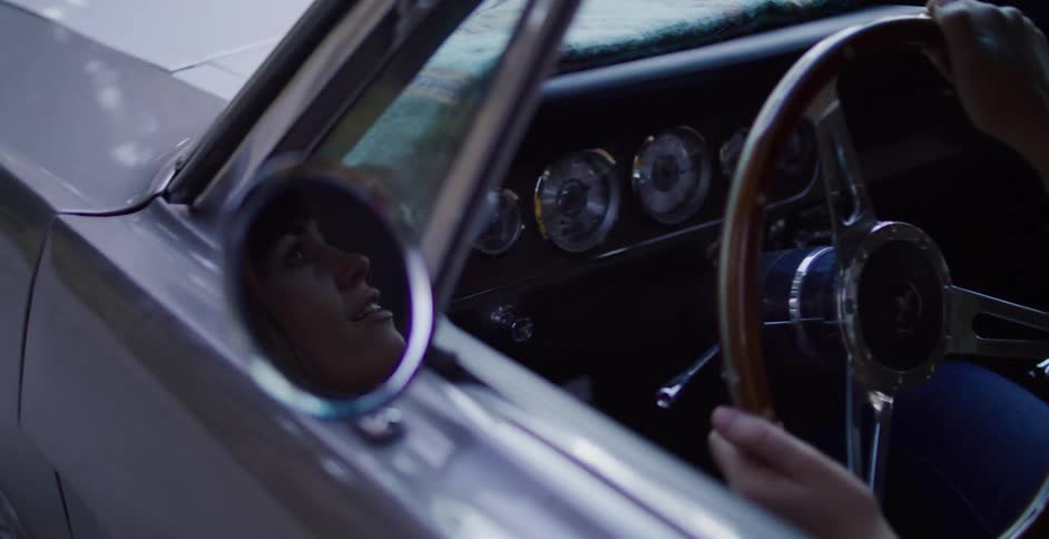 A woman's reflection is seen in the side mirror as she sits behind the steering wheel of a classic car