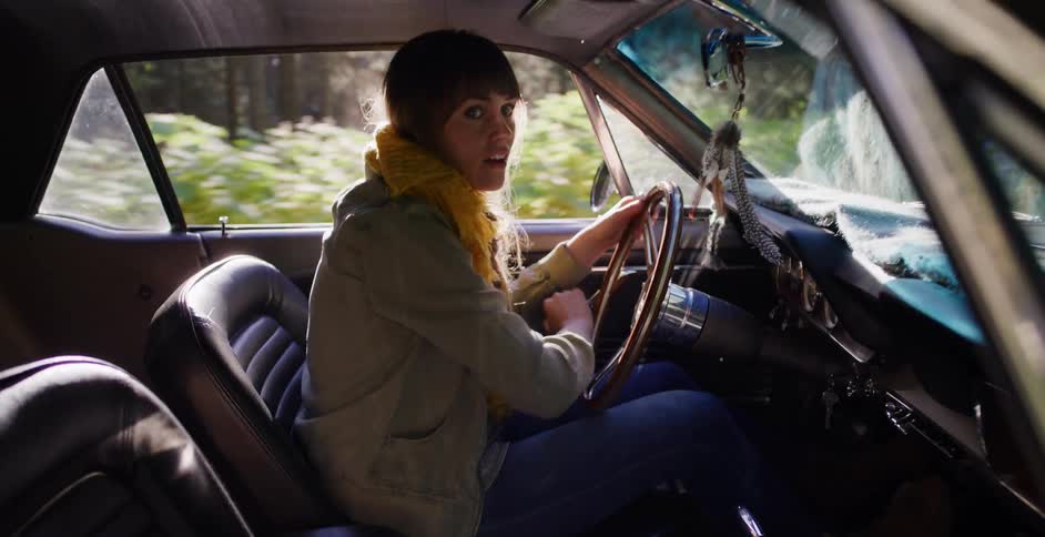 A young woman driving a classic car through a sun-dappled forest road looks back at the passenger seat in surprise