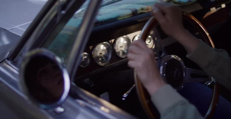 A woman's face is reflected in the side mirror of a classic car as her hands grip the wooden steering wheel