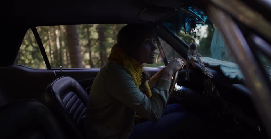 A young woman with a concerned expression drives her vintage muscle car through a forest as the light shifts around her