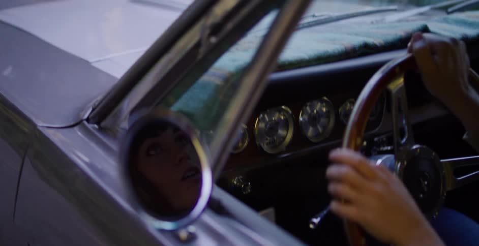A woman's face is reflected in the side mirror of a classic car as she drives, her expression shifting with the changing light