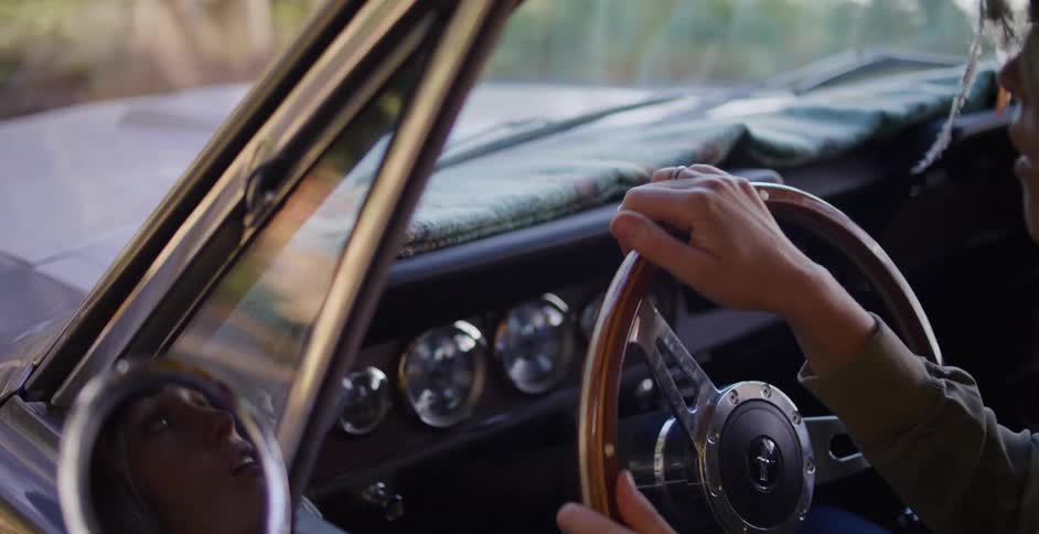 A woman's hands grip the wooden steering wheel as she drives a classic convertible car through a sunlit forest