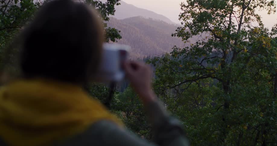 A person sips a warm beverage from a mug while peacefully watching the sun rise over a hazy mountain forest