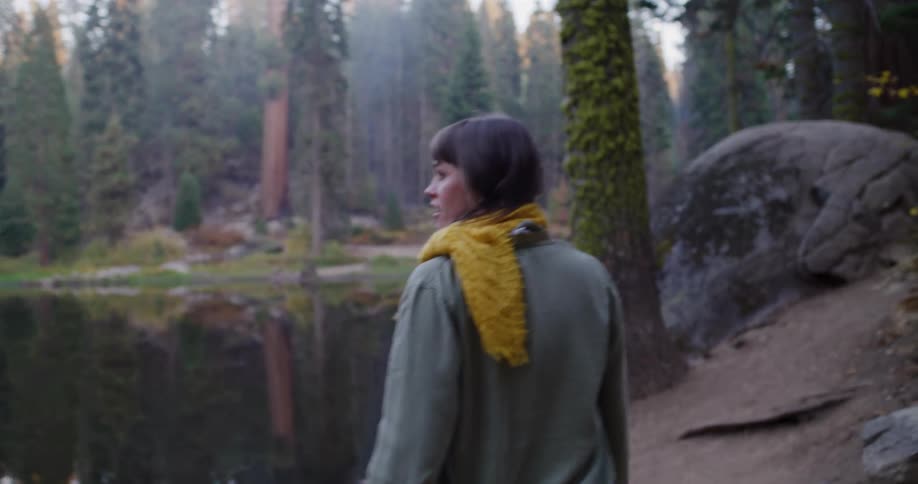 A woman with a yellow scarf walks on a trail alongside a serene lake in a dense forest