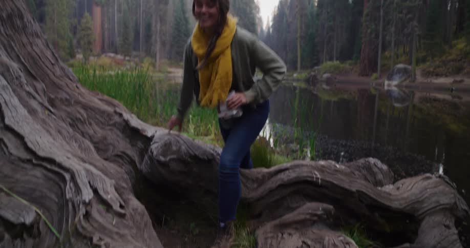 A smiling woman in a yellow scarf carefully hikes over giant tree roots next to a tranquil forest lake