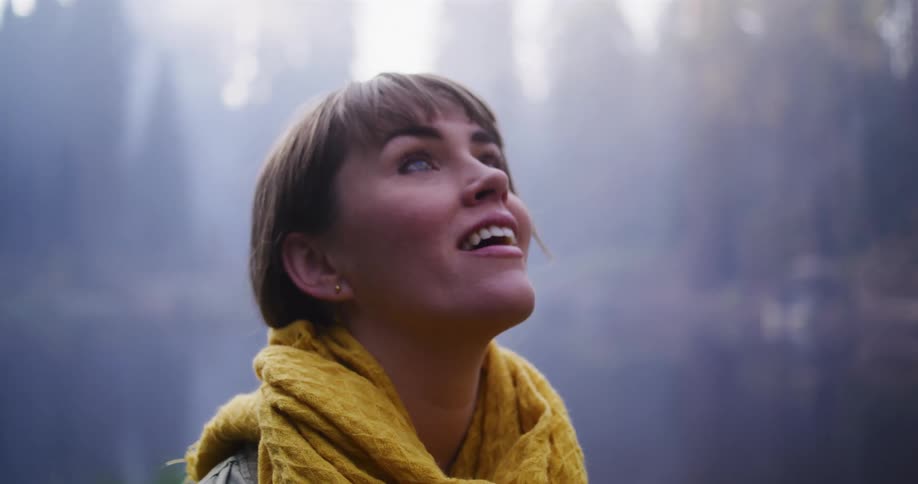 Close-up of a woman's face filled with wonder as she looks up at the sunlight filtering through a misty forest