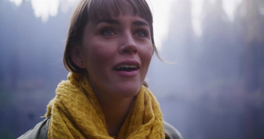 A woman with a yellow scarf gazes up in wonder and awe while standing in a misty, peaceful forest setting